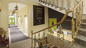 Eine Treppe mit aufwendigen goldfarbenen Geländern, die zu einer Zwischenebene führt. Ein Schild an der Wand zeigt den Schriftzug "SUPER SUPER ZOYPER SUPER SUPER".