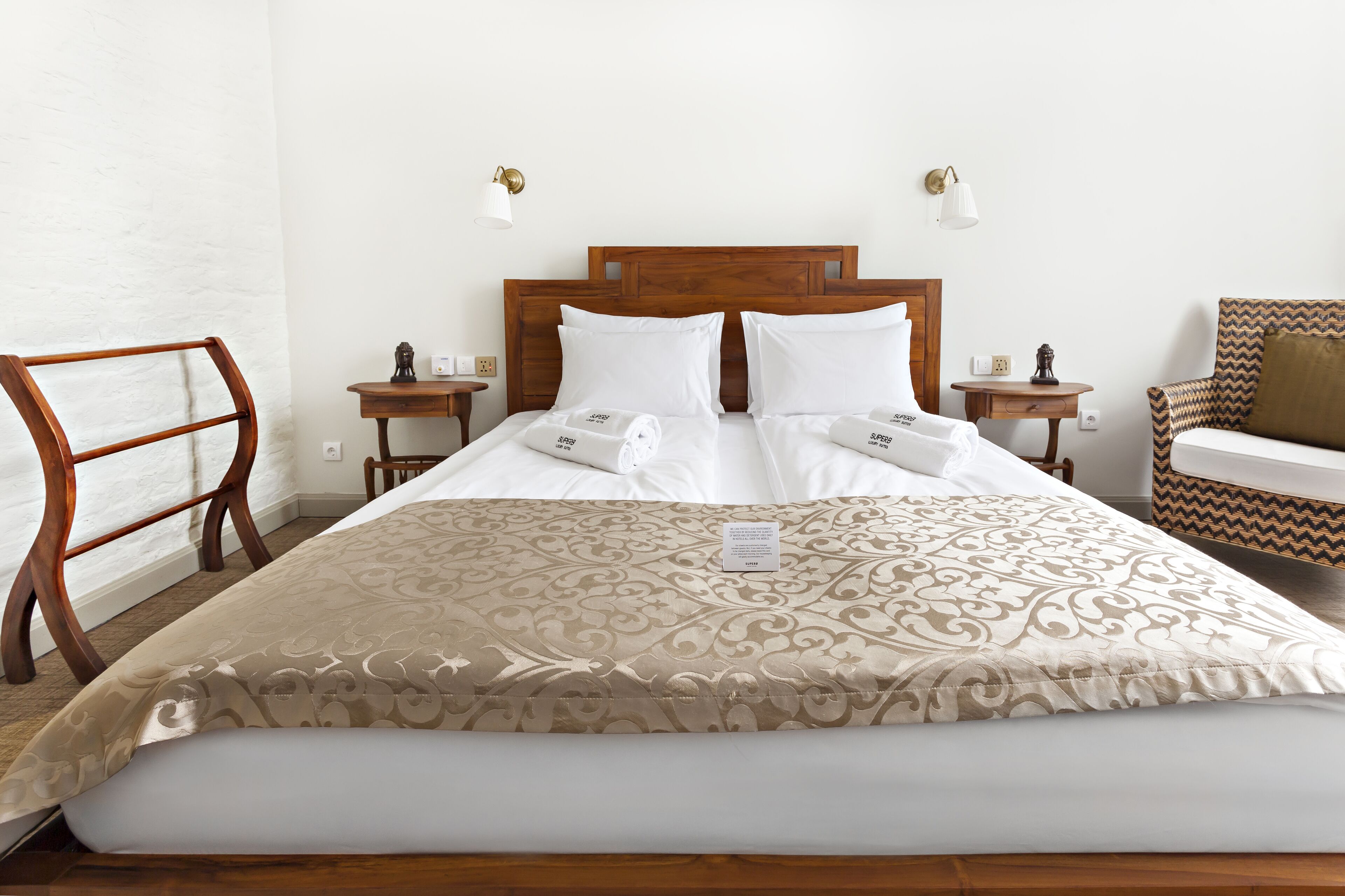 A neatly made bed with a patterned bedspread, two bedside tables with lamps, a wooden chair, and a sofa with a patterned cushion.