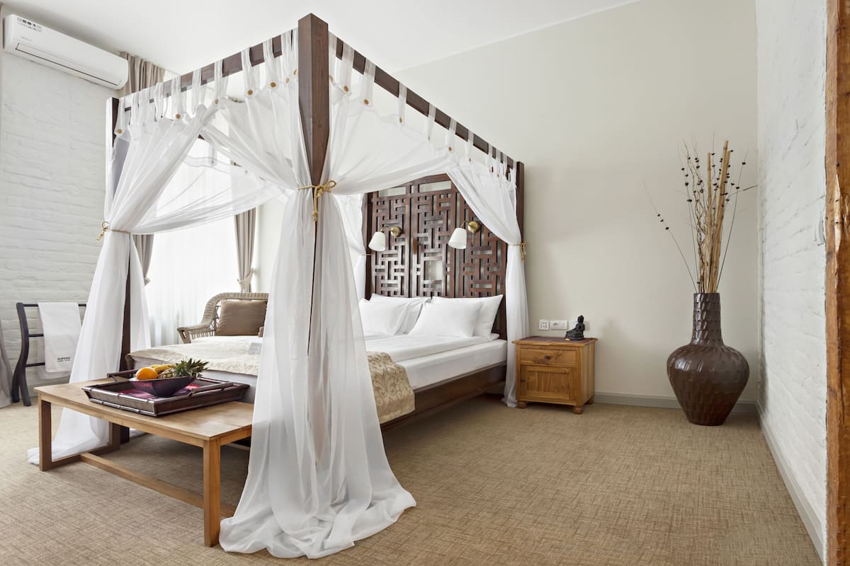 A bedroom with a canopy bed, bedside table with fruit, a nightstand, a large vase, and a decorative wall panel.