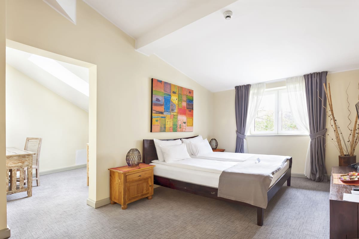 A bedroom with a bed, a desk, a chair, a painting, and a window with curtains.