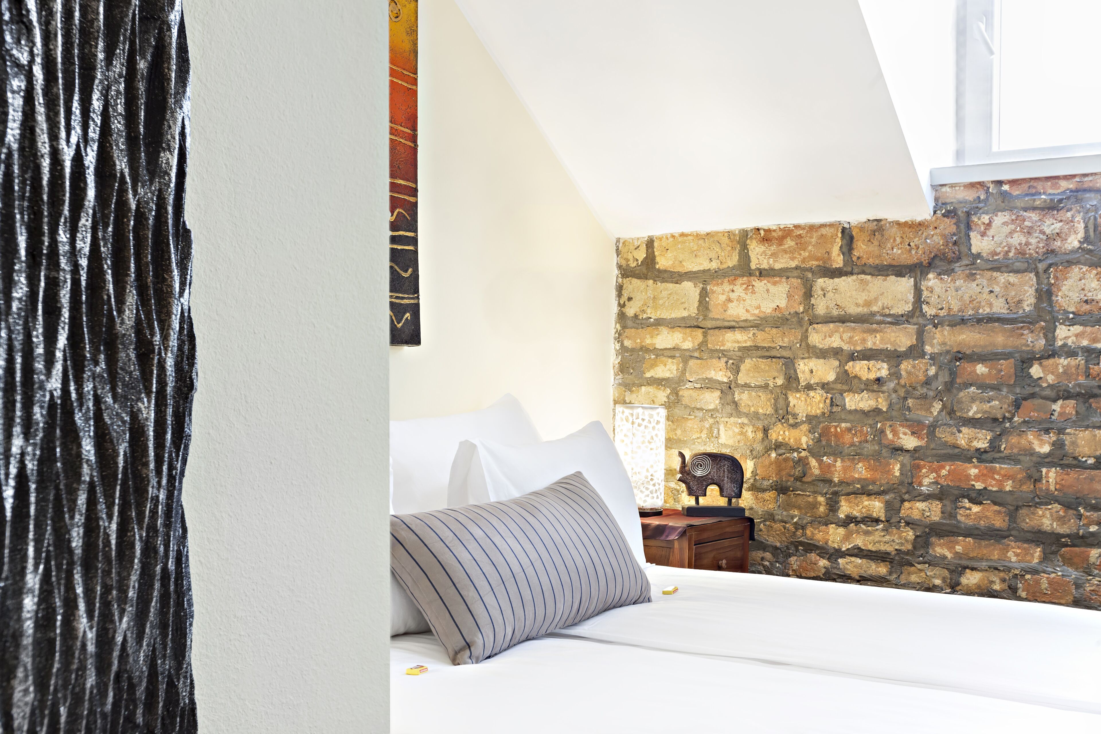Una camera da letto con parete in mattoni, un letto con biancheria bianca, un cuscino a righe e un piccolo comodino in legno con una lampada.