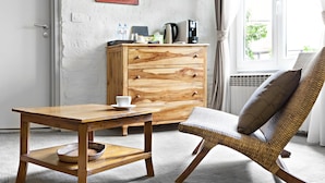 Ein Zimmer mit einem hölzernen Schmuckkästchen, einem Korbstuhl, einem Holztisch mit Tasse und einem Bild an der Wand.