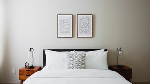 Ein ordentlich bezogenes Bett mit weißen Leinen, einem schwarzen Kopfteil und einem Kissen mit geometrischem Muster. Über dem Bett hängen zwei gerahmte abstrakte Kunstwerke.