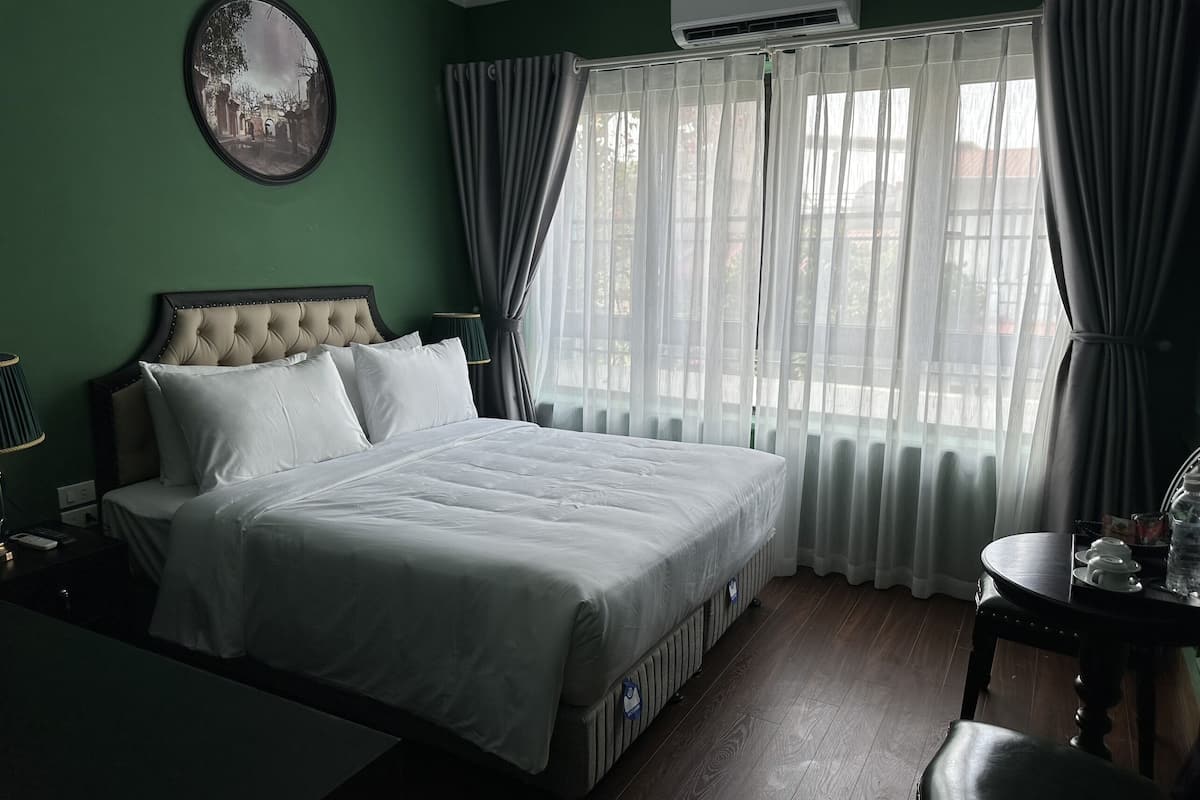 A hotel room with a large bed, a bedside table, a chair, a small table with cups and a bottle, and a view of the outside through sheer curtains.
