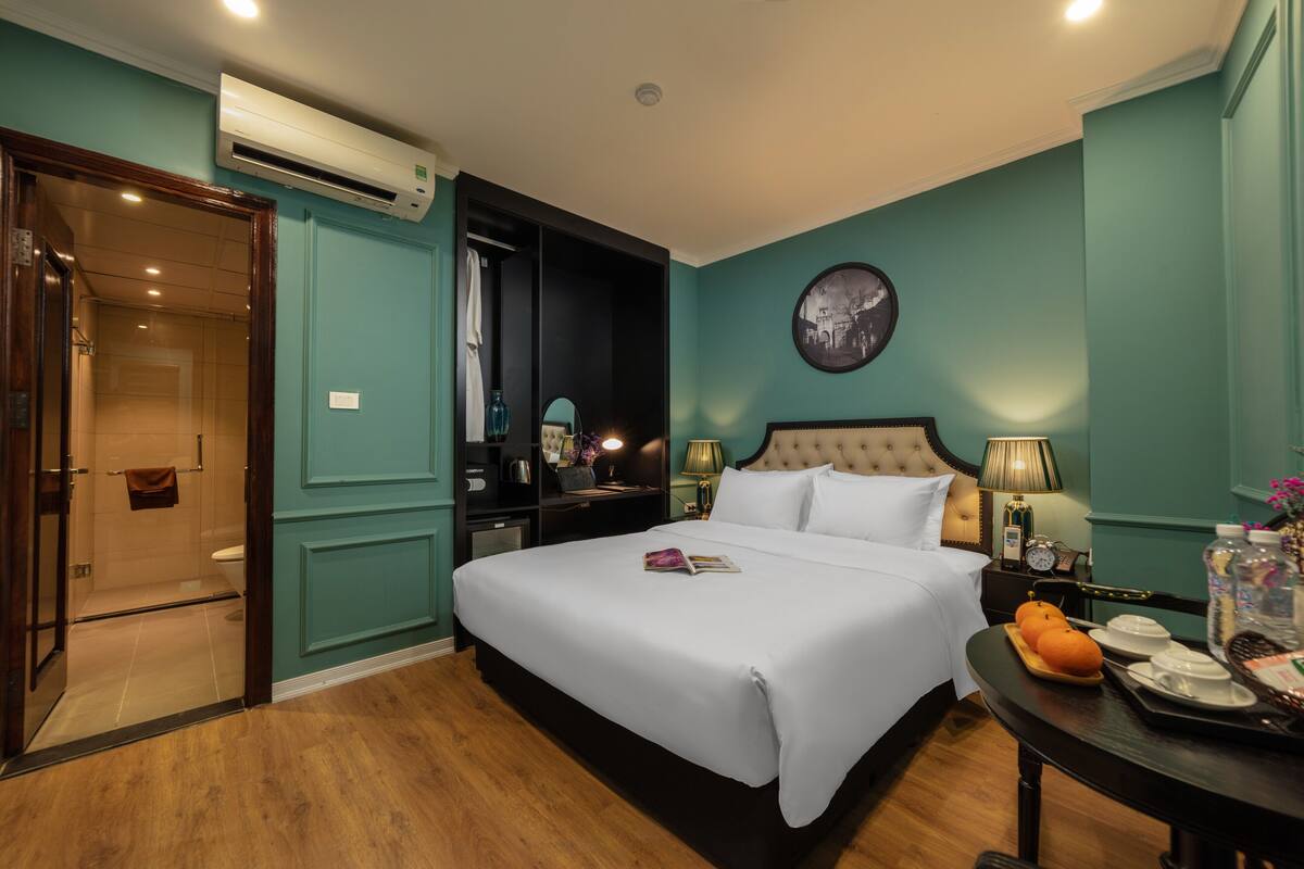 A hotel room with a large bed, a bedside table with a lamp, a small table with oranges and a tray, and an air conditioning unit on the wall.