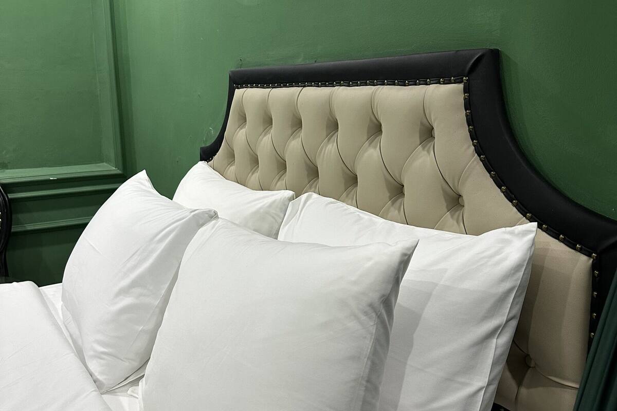 A bed with white linens and a tufted headboard, set against a green wall with a round mirror reflecting a street scene.