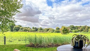 Een terras met een ronde tafel en stoelen, met uitzicht op een groen veld en bomen onder een gedeeltelijk bewolkte lucht.