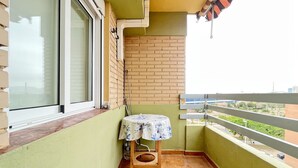 Un petit balcon avec un sol carrelé, une table recouverte d’une nappe à motifs floraux et une vue sur le paysage urbain.
