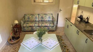 Ein kleiner, gemütlicher Wohnbereich mit einer blumigen Couch, einem Esstisch mit Platzdeckchen, einem kleinen Beistelltisch mit Lampe und einer Küchenzeile mit Spüle und Schränken.