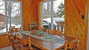 A wooden dining room with a large table and chairs, large windows with a snowy view, and a small side table with glassware.