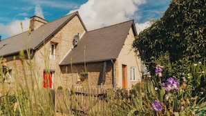 A stone house with a sloped roof, a wooden fence, and a garden with purple flowers. - Gîte Charmant à la Campagne (Chérencé-le-Héron)