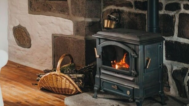 A stone fireplace with a wood-burning stove, a basket, and a kettle on the mantel. - Gîte Charmant à la Campagne (Chérencé-le-Héron)