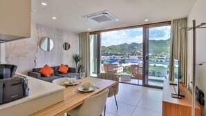 A modern hotel room with a dining area, a sofa with orange cushions, a coffee machine, and a view of a coastal town. - A116 - Immersive Simpson Bay Lagoon View (Sint Maarten)