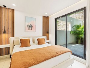A modern bedroom with a large bed, a wooden accent wall, a framed picture, and a sliding glass door leading to a balcony with plants. - A101 - Outstanding Lagoon View & Garden (Sint Maarten)