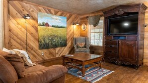 Een kamer met houten lambrisering, een groot schilderij, een televisietoestel, een geruit fauteuil en een houten kast.