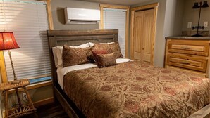 Ein Schlafzimmer mit einem hölzernen Bett, gemusterten Betttextilien, einer Nachttischlampe, einer Holzkommode und einem Fenster mit Jalousien.