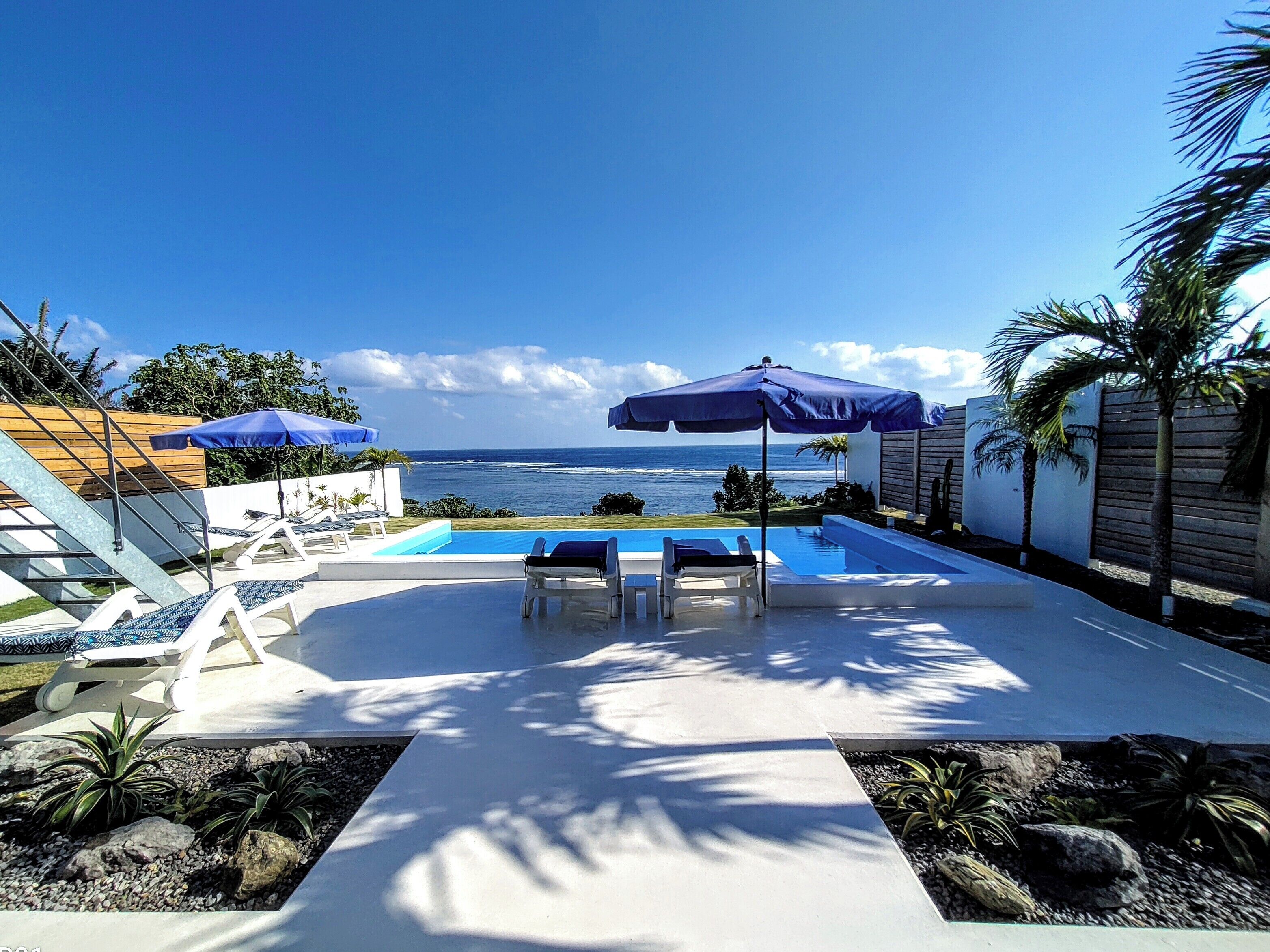Zona de piscina con sillones de descanso y sombrillas, vista al océano y un jardín cuidado con rocas y plantas.