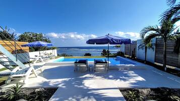 Zona de piscina con sillones de descanso y sombrillas, vista al océano y un jardín cuidado con rocas y plantas.