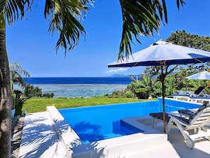 A swimming pool with lounge chairs and umbrellas, surrounded by palm trees and a view of the ocean. - Ocean View Ishigaki Sora and Umi (Ishigaki)