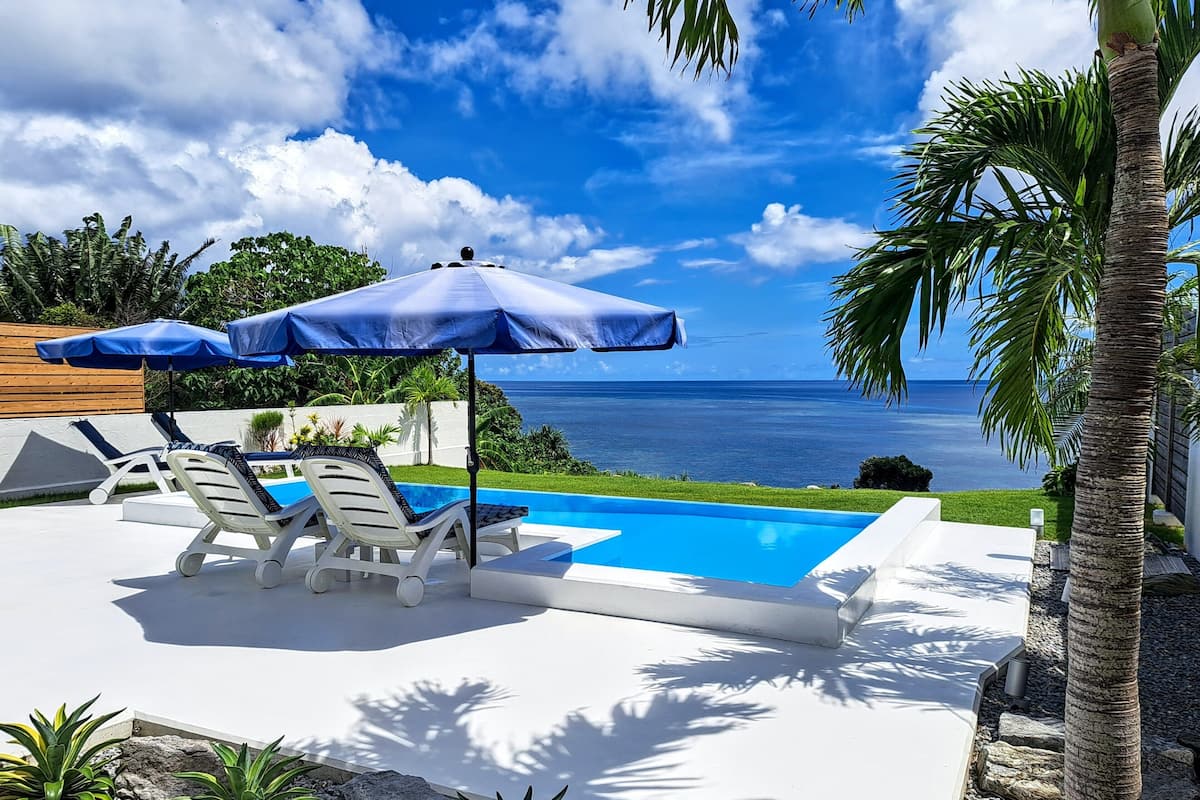 A pool area with lounge chairs and umbrellas, a view of the ocean, and a well-maintained garden.