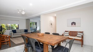 Uma sala de estar ampla com uma mesa de jantar de madeira, cadeiras estofadas cinzas, um sofá cinza e um banco de madeira. Há um ventilador de teto, uma janela grande e uma porta que dá para uma varanda.