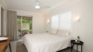 Quarto com cama, ventoinha no teto, janela com persianas, uma pequena mesa com uma planta e vista para um pátio com cadeiras e mesa.