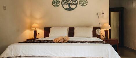 A neatly made bed with white linens and two rolled towels, a wooden headboard, two bedside lamps, and a wall decoration of circular tree designs.