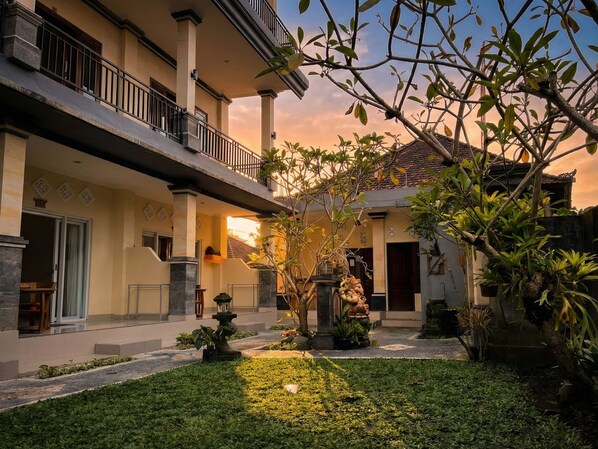 Un bâtiment à deux étages doté d’un balcon, un jardin avec une fontaine et un coucher de soleil en arrière-plan.
