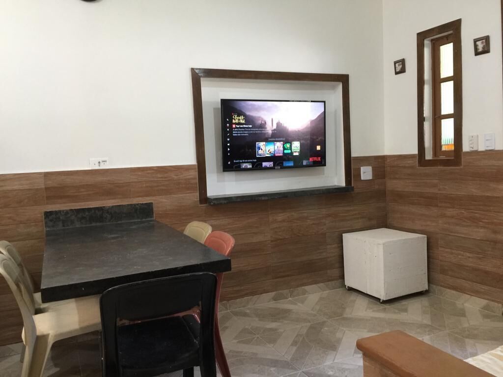 Quarto com televisão na parede, uma mesa, cadeiras e um pequeno armário branco.