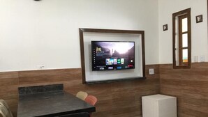 Una habitación con una televisión montada en la pared, una mesa, sillas y un pequeño armario blanco.