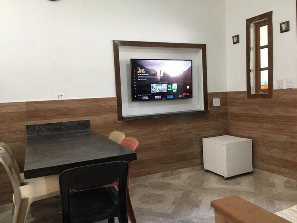 A room with a TV mounted on the wall, a table, chairs, and a small white cabinet.