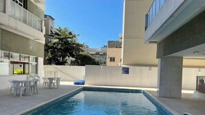 A rectangular swimming pool with a tiled edge, surrounded by a patio area with white plastic chairs and a small table. - Precioso Apartamento con Terraza y Piscina (Ipanema Rio de Janeiro)