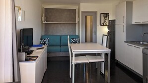 A compact kitchen with a dining table, a sofa, and a refrigerator. - Studio Heliopolis K Int. Naturist Village AC/Park (Agde)