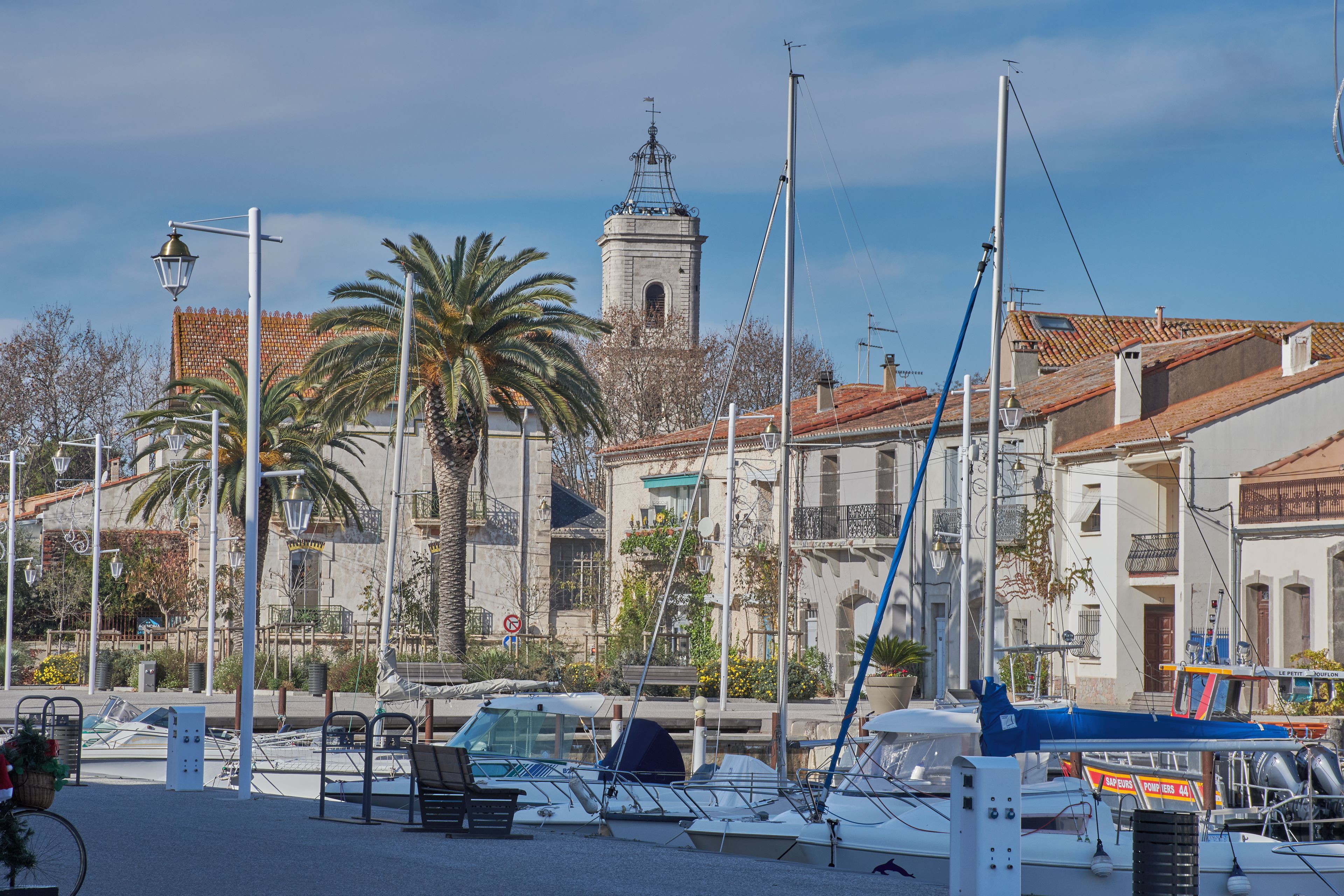 Un port de plaisance avec des bateaux amarrés, un palmier et des bâtiments aux toits recouverts de tuiles rouges.