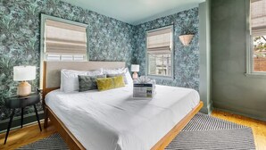 Un grand lit avec une literie blanche, une tête de lit en bois et des coussins décoratifs. Il y a deux fenêtres avec des stores, un ventilateur de plafond et une petite table avec une lampe.