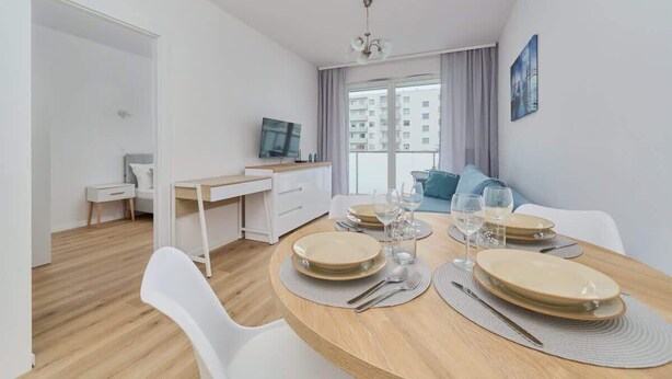 Ein modernes, helles Zimmer mit Esstisch für vier Personen, einem Fernseher und Blick auf eine Stadtlandschaft durch große Fenster.