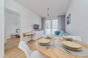 Una habitación moderna y luminosa con una mesa de comedor preparada para cuatro personas, un televisor y vistas a un paisaje urbano a través de grandes ventanales.