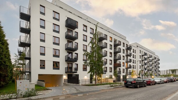 Un immeuble résidentiel moderne à plusieurs étages, doté de balcons, d’une entrée de garage et d’une rangée de voitures garées.