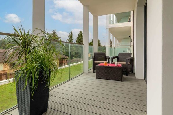 A balcony with a potted plant, wicker furniture, and a glass railing overlooking a green landscape.
