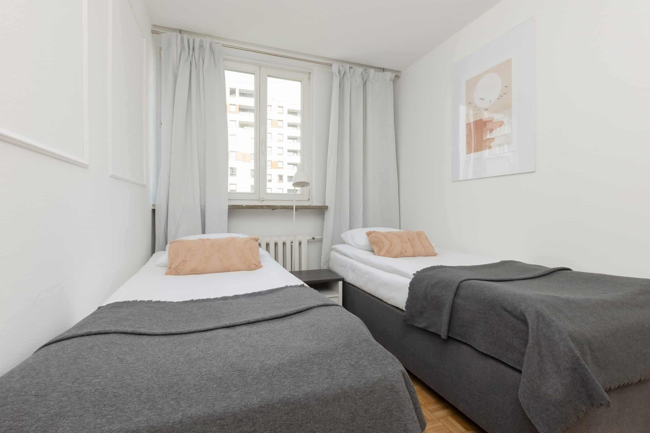 A small, modern room with two beds, a window with curtains, and a wall-mounted artwork.