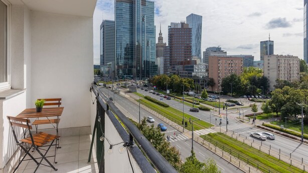 A balcony with a table and chairs, overlooking a cityscape with modern buildings and a busy street. - Spacious Apartment in the Center | Balcony and Workspace (Warszawa)