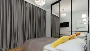 Une chambre moderne avec un grand lit, des rideaux gris, un lustre, un téléviseur à écran plat et un miroir avec un cadre noir.