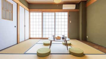 Ein Raum mit Tatami-Matten, einem niedrigen Tisch, Kissen, Schiebetüren und einem Deckenventilator.
