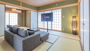 Un salon de style japonais traditionnel, avec un sol recouvert de tatamis, un plafond bas en bois et un téléviseur fixé au mur.