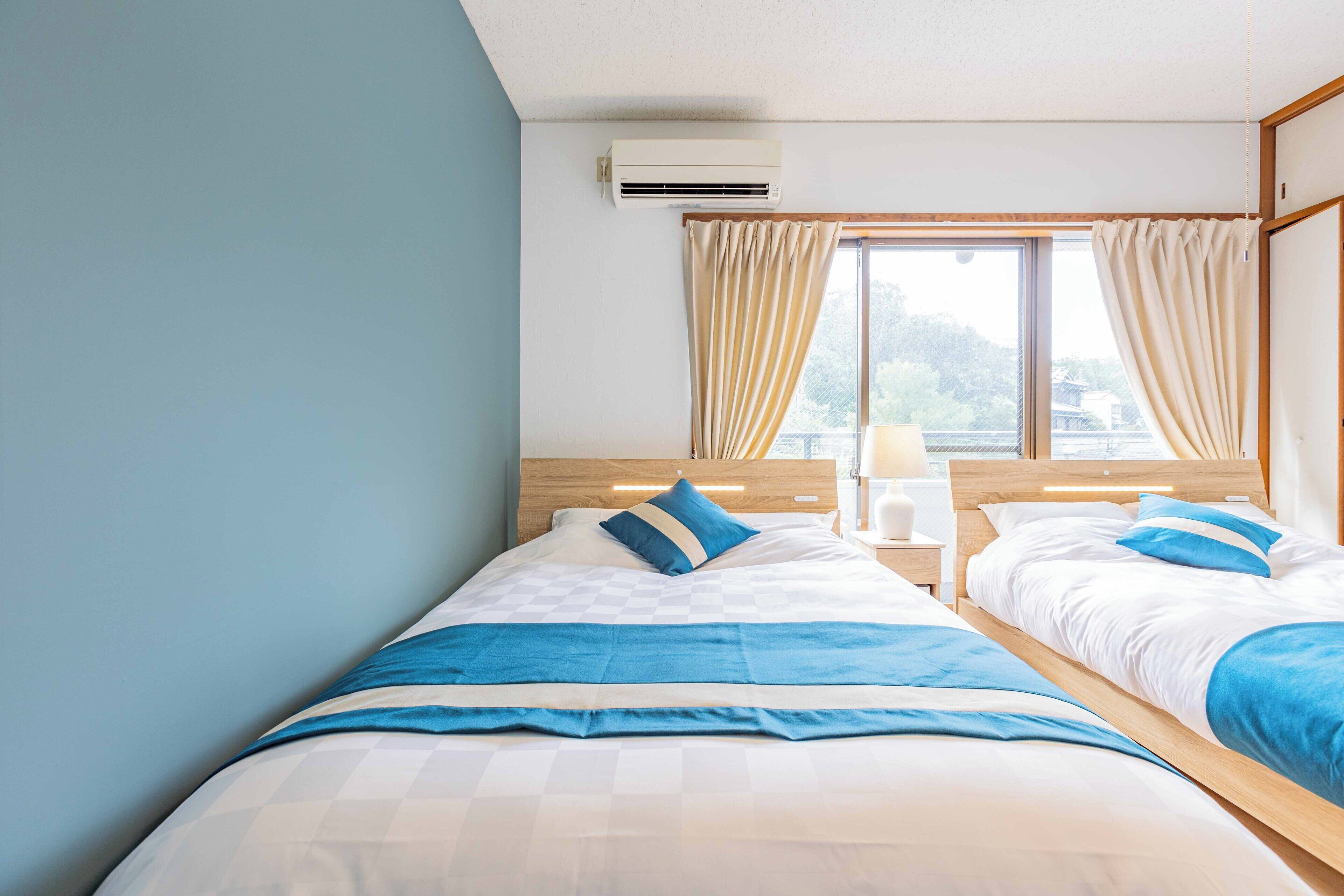 A hotel room with two beds, a wooden headboard, a nightstand with a lamp, and a window with curtains.