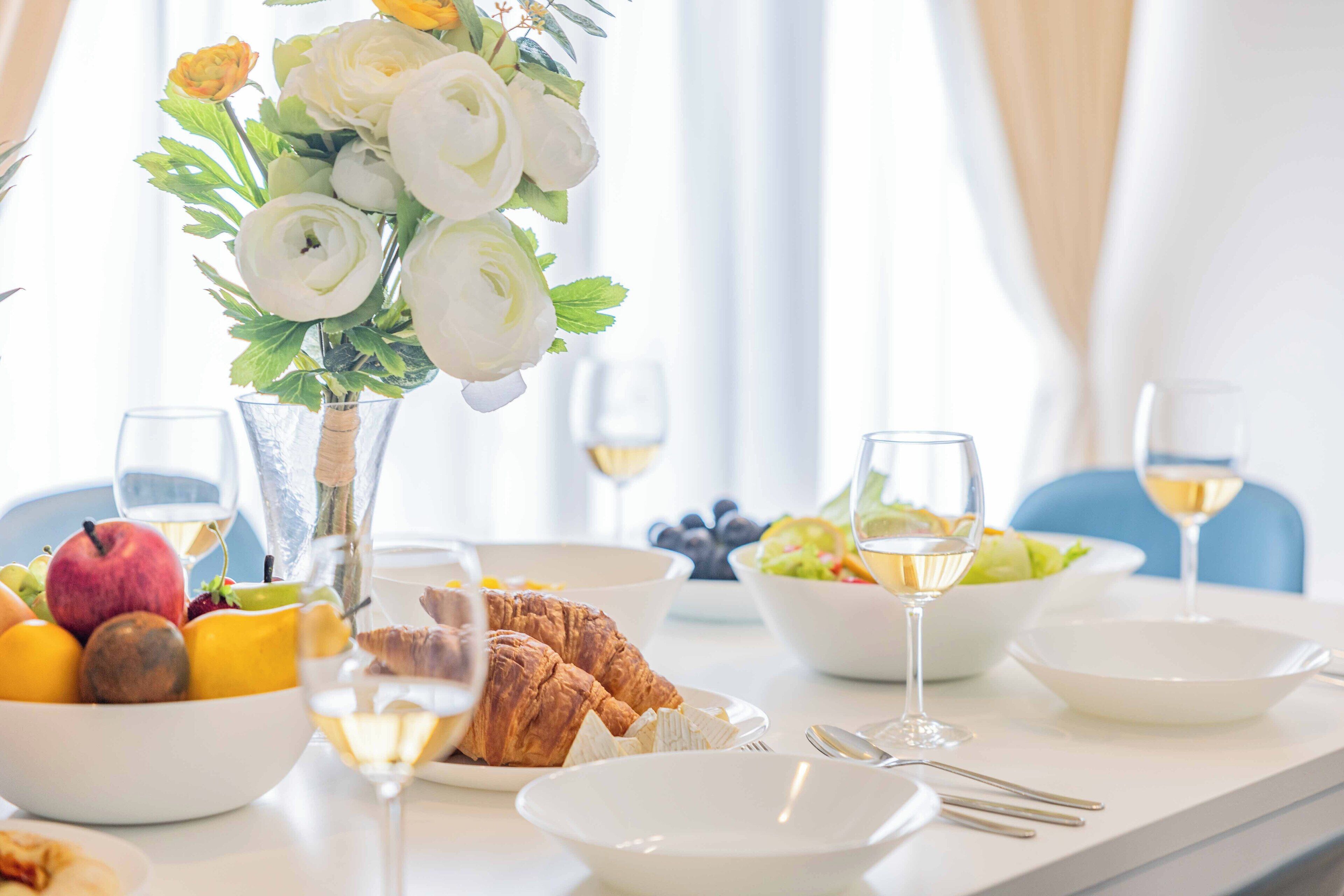 Une table à manger avec un panier de fruits, des croissants, des verres à vin et un vase de fleurs.