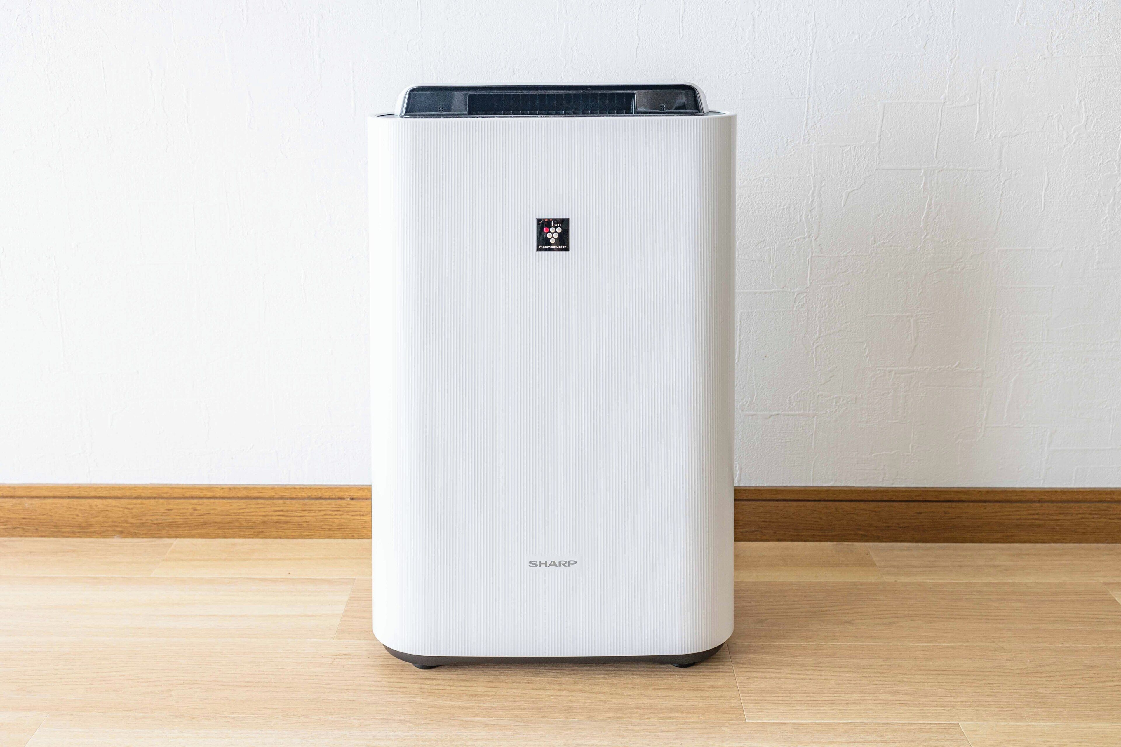 Un purificador de aire blanco, con textura vertical, de forma rectangular, con un panel de control en la parte superior y el nombre de la marca 'SHARP' en la parte inferior.
