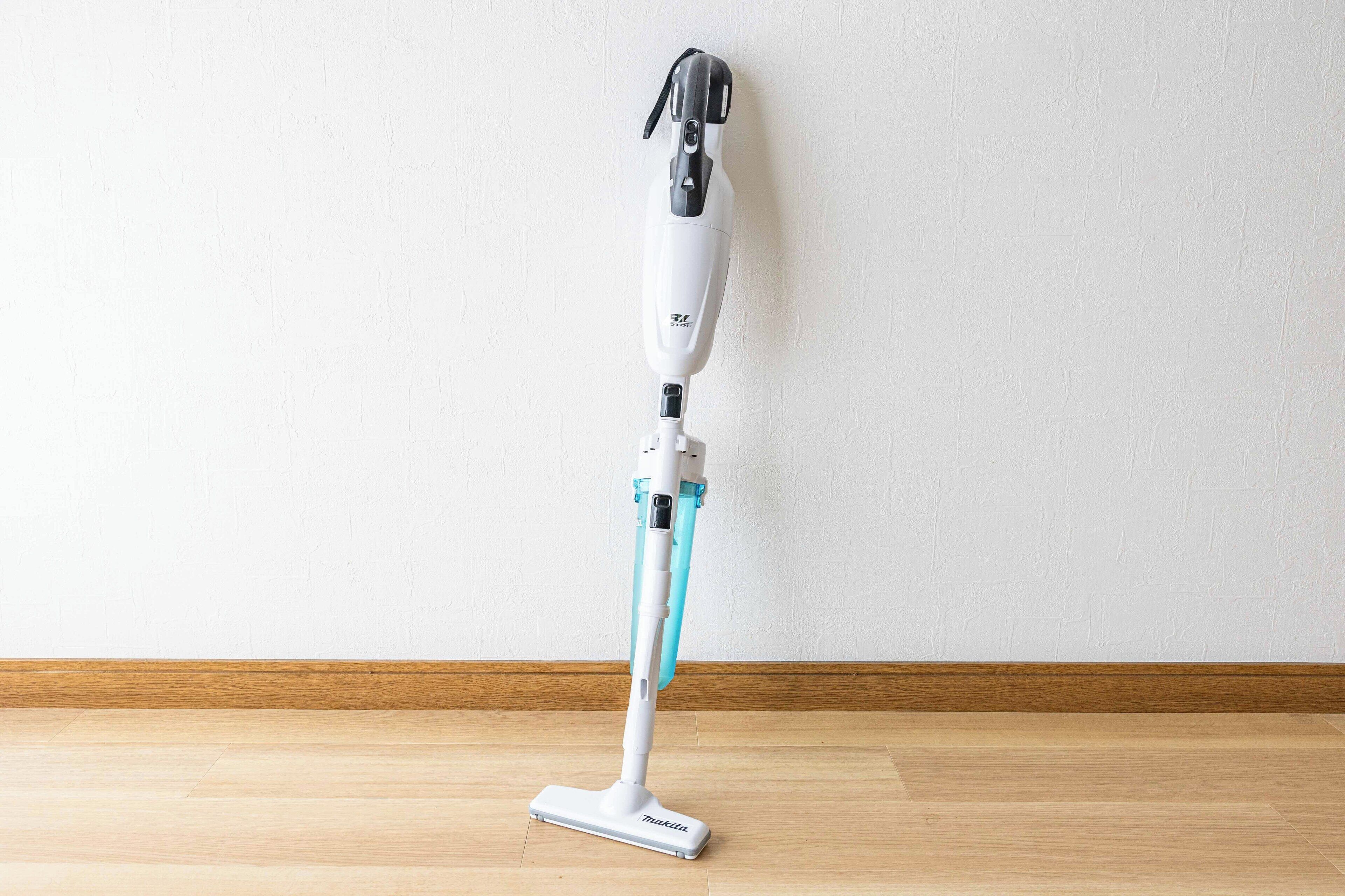 Een stofzuiger met een lange steel en een dweilopzetstuk, staand op een houten vloer tegen een witte muur.