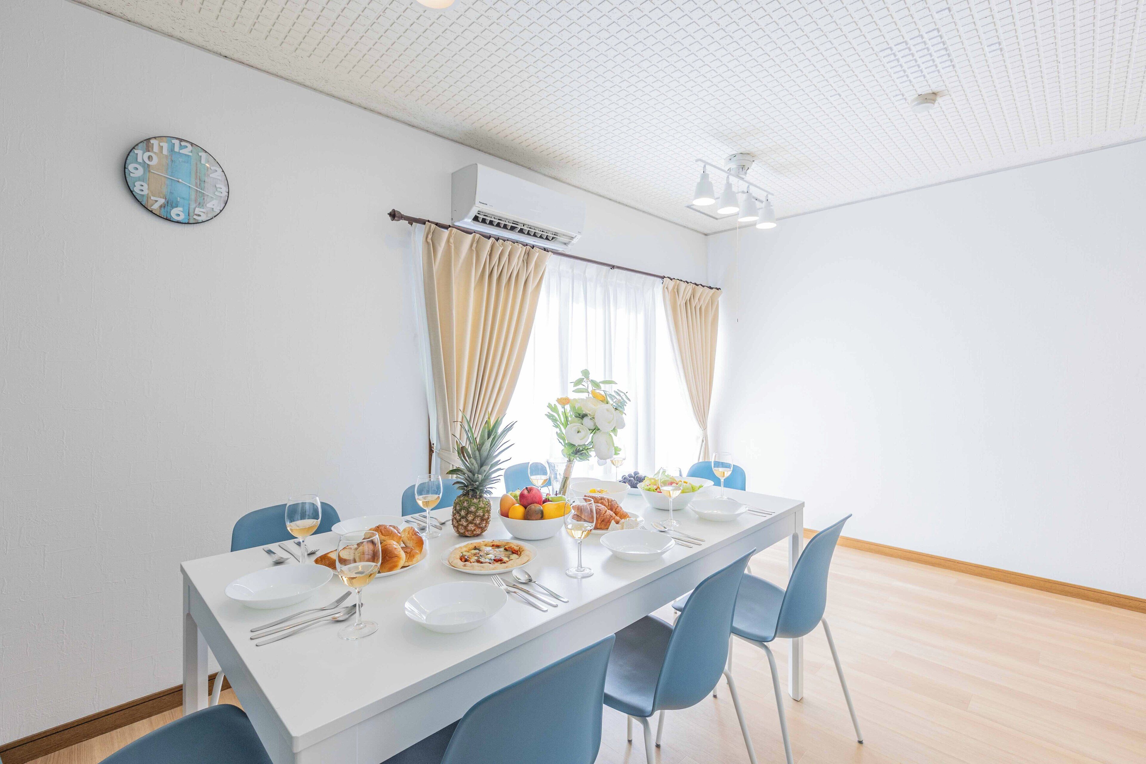 Un comedor con una larga mesa dispuesta para cuatro personas, paredes blancas, un techo con diseño de cuadrícula y un reloj en la pared.