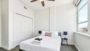 A hotel room with a large bed, a ceiling fan, a window with a view, a wardrobe, and a bedside table with a lamp. - Modern Downtown High-Rise Apartment (Baton Rouge)